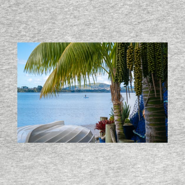 Tropical garden frames view over white clinker dinghy to bay with stand-up paddle-boarder in distance. by brians101